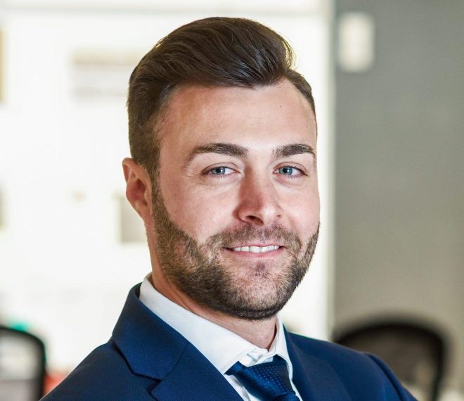 Caucasian businessman leader looking at camera in modern office with multi-ethnic businesspeople working at the background. Teamwork concept. Young man with beard wearing blue suit.